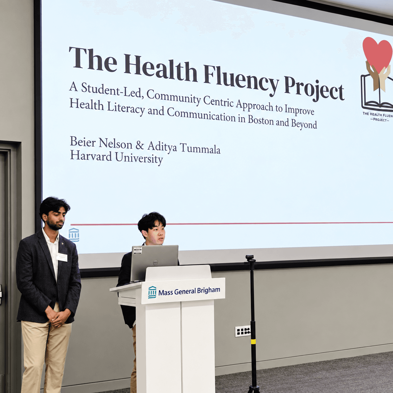 Aditya Tummala and Beier Nelson presenting The Health Fluency Project beside a slide introducing its student-led, community-centric approach.
